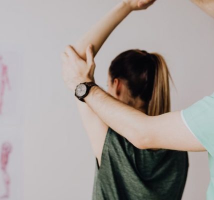 Bearded chiropractor in eyeglasses and wristwatch examining arm of anonymous female in casual clothes with raised hand in doctor office in hospital