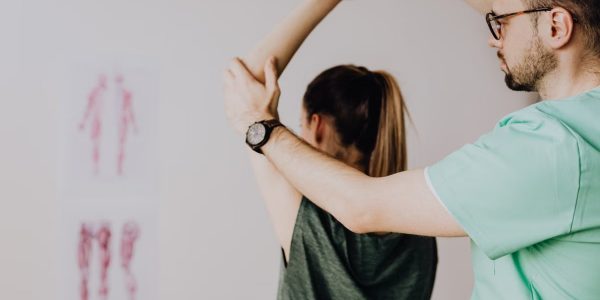 Bearded chiropractor in eyeglasses and wristwatch examining arm of anonymous female in casual clothes with raised hand in doctor office in hospital