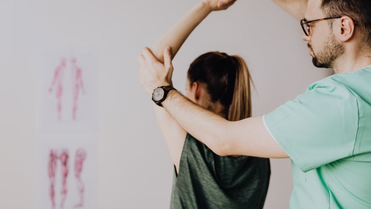 Bearded chiropractor in eyeglasses and wristwatch examining arm of anonymous female in casual clothes with raised hand in doctor office in hospital